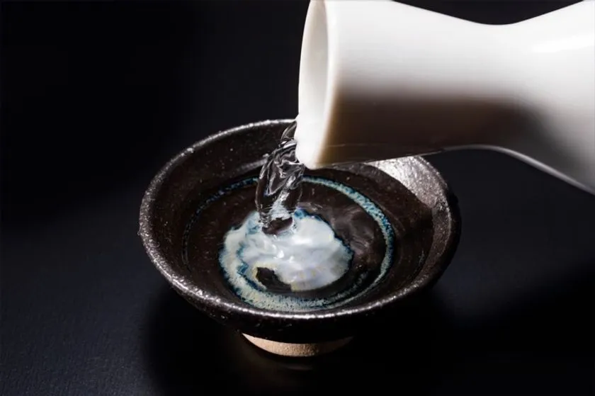 白い徳利から黒釉の盃に日本酒を注ぐ瞬間をクローズアップした暗背景のシズル写真