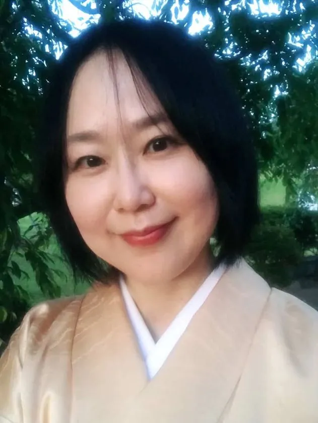 薄金色の着物と白い半衿を着用した黒髪ショートの女性が屋外の緑の木々を背景に正面を向いて微笑む人物写真