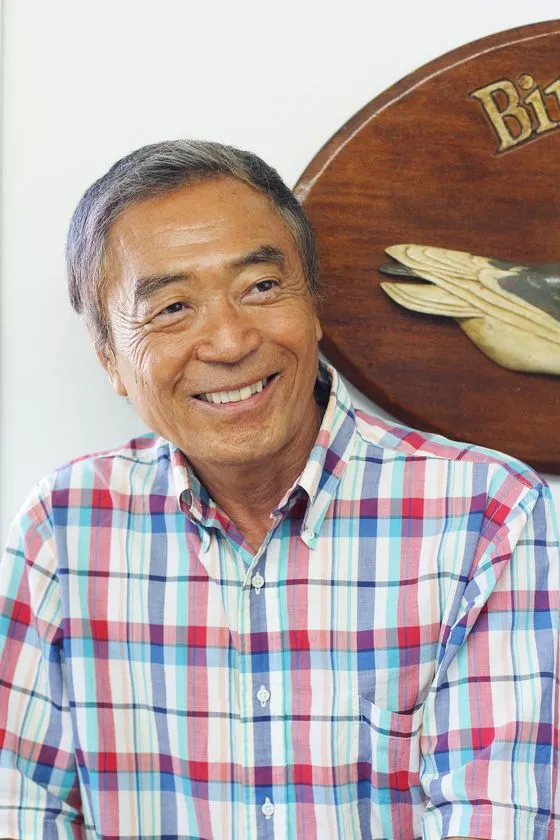 チェック柄シャツを着た浅井愼平本人の顔写真。木製の円形プレートを背景に正面向きで微笑む上半身ポートレート。