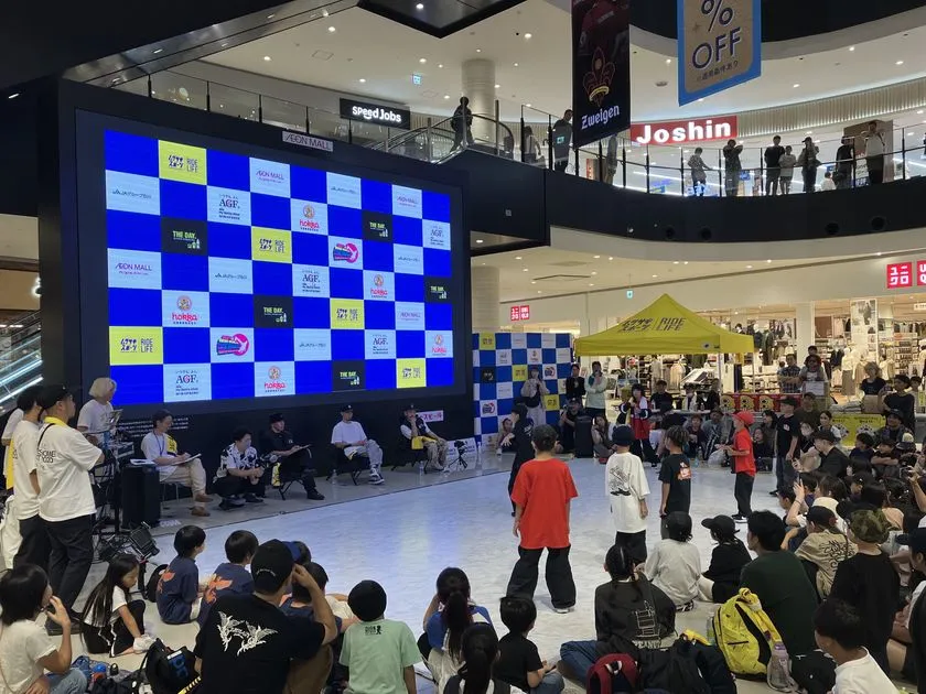 イオンモール館内の吹き抜けコートで開催されたブレイキン大会の競技風景。参加選手2名がフロアで向き合い、審査員席・大型スポンサーLEDビジョン・多数の観客が写る
