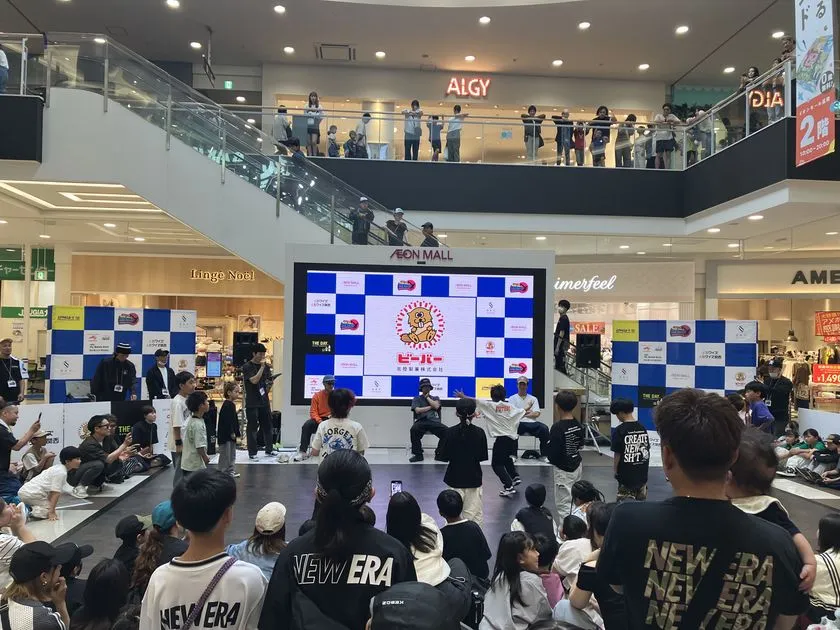 イオンモール館内コートでのブレイキン大会で複数選手がフロアに集まりパフォーマンス中の場面。スポンサーロゴ入りLEDビジョンと2階ギャラリーを含む多数の観衆が囲む