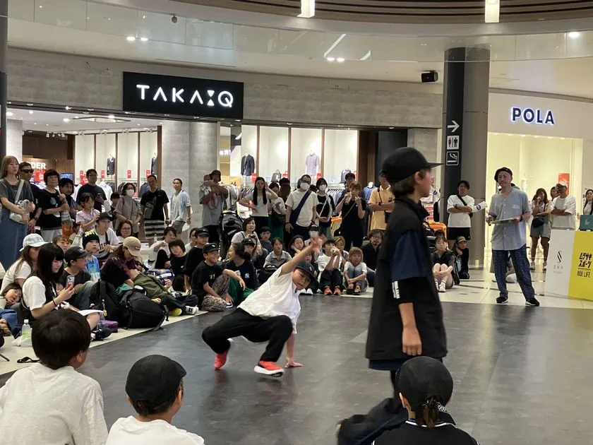 イオンモール館内(TAKA-Q・POLA店舗前)でブレイクダンス大会が開催され、白Tシャツ姿の子どもダンサーが低い姿勢でステップを踏み、周囲に多数の観客が座り込んで観戦している