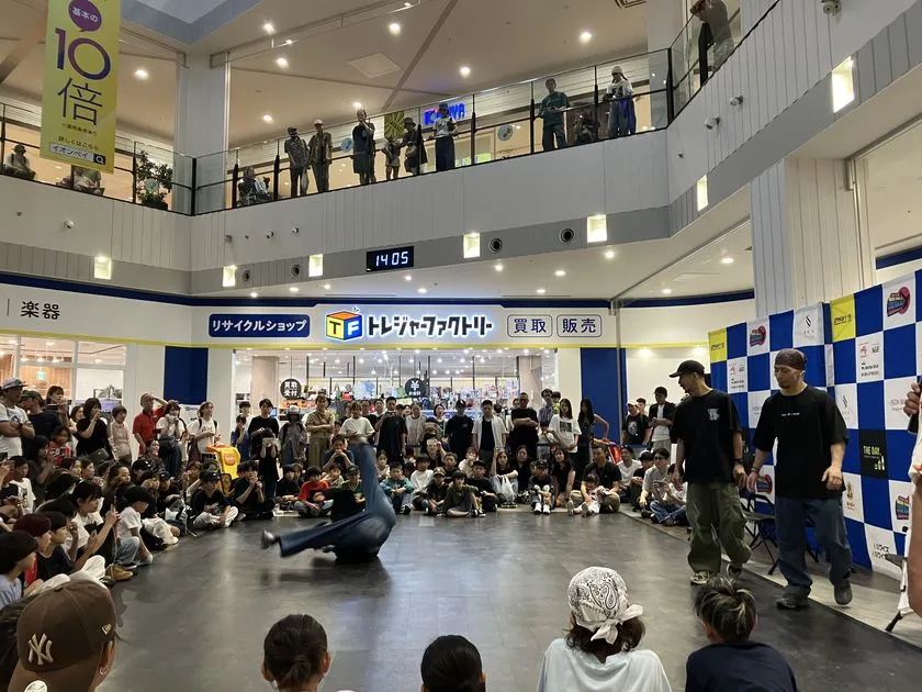 イオンモール吹き抜けコートでブレイクダンス大会の対戦シーンを撮影し、1名がフロアムーブ中、ジャッジと選手が円形に並び2階からも観客が見下ろす様子と時刻14:05の電光掲示板が写っている