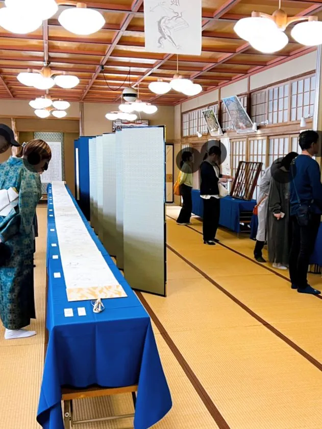 和室の大広間に長机を並べて手漉き和紙絵巻を展示し、来場者数名が間近で鑑賞している会場内の実景写真。畳敷き・格天井・障子という伝統的な建築空間での展示の様子がわかる