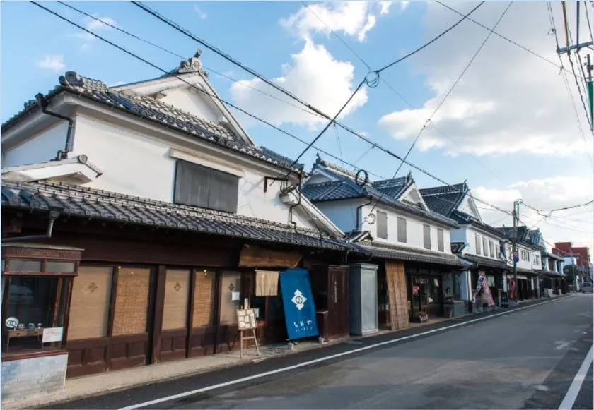 福岡・八女の伝統的な商家の町並みを撮影した外観写真。瓦屋根の白壁蔵造り建物が通り沿いに連続し、藍色の暖簾を掛けた店舗が展示会場周辺の雰囲気を伝える