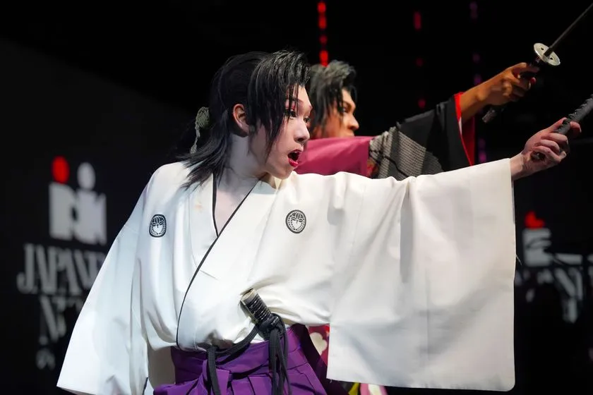 白い紋付き羽織と紫袴を着けた女性パフォーマーが刀を前方に突き出し口を開いて演じる殺陣・武道パフォーマンスの舞台写真