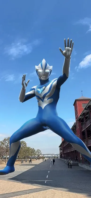 横浜赤レンガ倉庫前の広場に巨大なウルトラマン（青と銀の配色）がARで出現し両手を広げたポーズで降り立つ体験イメージ写真