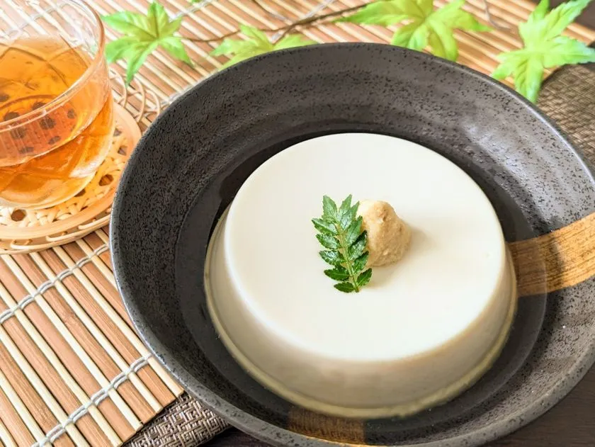 北海道山わさび豆腐の盛り付け完成写真。黒い陶器の深皿に白いおぼろ豆腐をドーム型でよそい、山わさびすりおろしと木の芽をトッピング。竹製ランチョンマットと麦茶のグラスを添えた和食スタイルの実食イメージ。