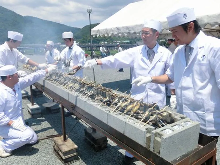 白衣・白帽の料理人複数名が嵐山を背景に屋外の長尺コンクリート製炭火台で串刺し若鮎を一列に並べて焼く若鮎試食コーナーの実写