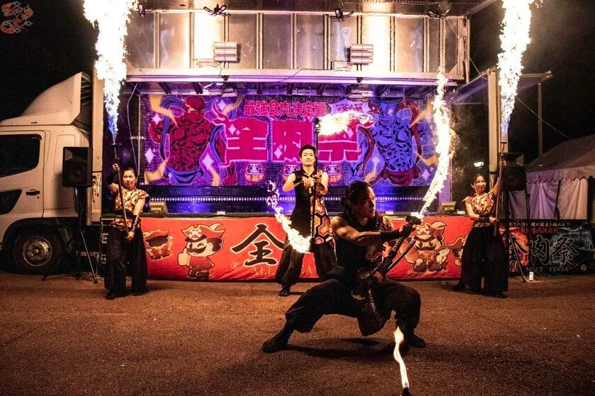 全肉祭会場での夜間ファイヤーパフォーマンス実演写真。全肉祭ロゴ入りトラックステージ前で火を操る男性パフォーマー1名と女性ダンサー2名が演技中の実写カット