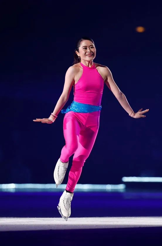 ピンクのノースリーブコスチュームにブルーのウエストリボンをつけた女性スケーターがリンク上でジャンプ直後の着氷姿勢をとるパフォーマンスシーン
