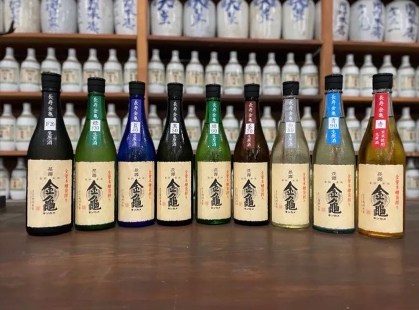 金亀酒造の長寿金亀シリーズ9本を精米歩合F20〜赤ラベル順に横一列で並べた全ラインナップ比較ショット。蔵内棚を背景に撮影
