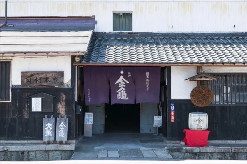 銘柄「金亀」の酒蔵・岡村本家の正面外観。紺色の暖簾と鏡割り用の酒樽が入口に並び、瓦屋根の白壁蔵が写る