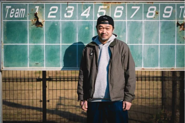 野球場のスコアボードを背景にキャップをかぶった男性が正面を向いて立つポートレート写真