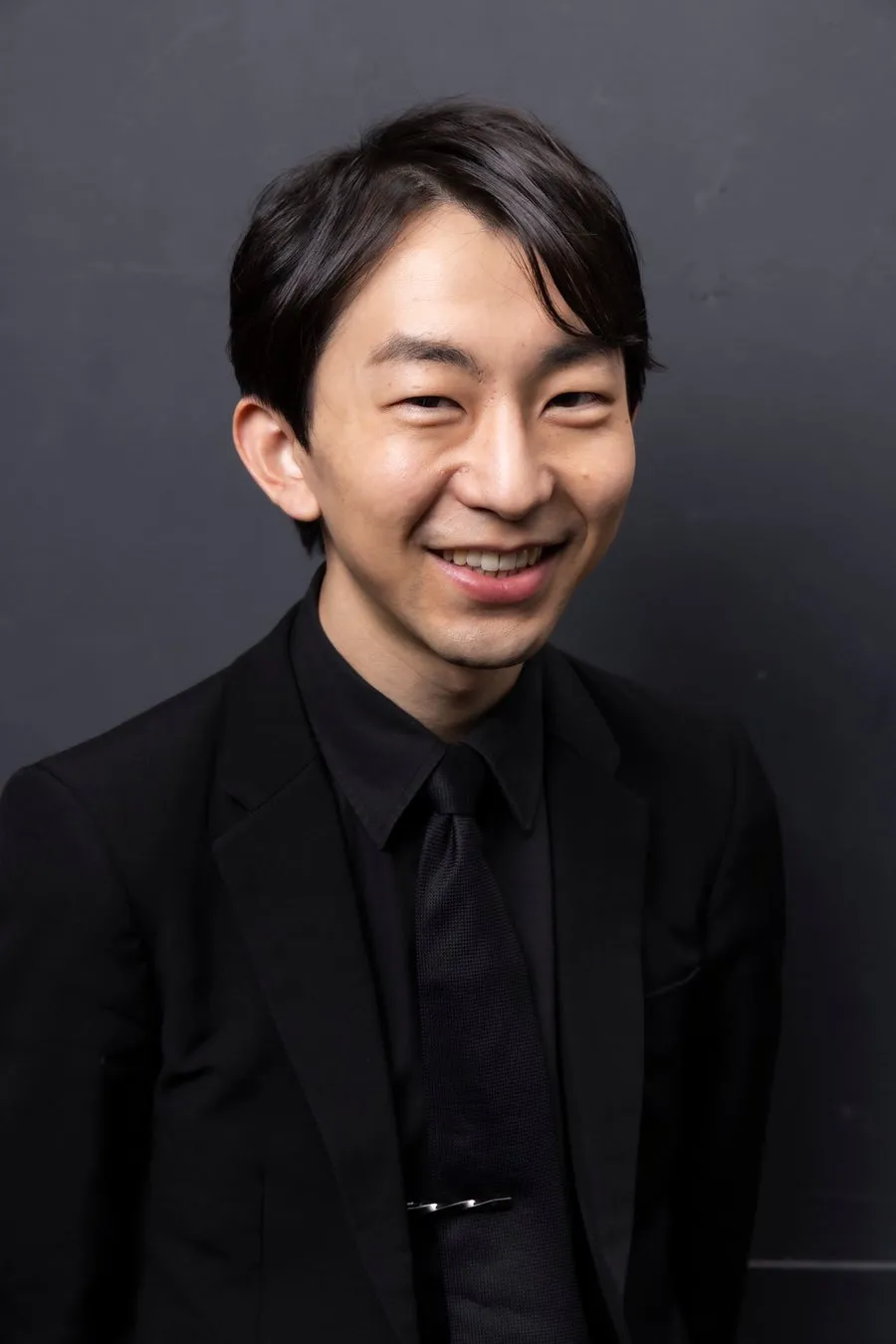 お笑い芸人・リップグリップ岩永圭吾さんの宣材写真風ポートレート。黒スーツ・黒シャツ・黒ネクタイ姿で笑顔を見せる男性の上半身が灰色背景に映る。