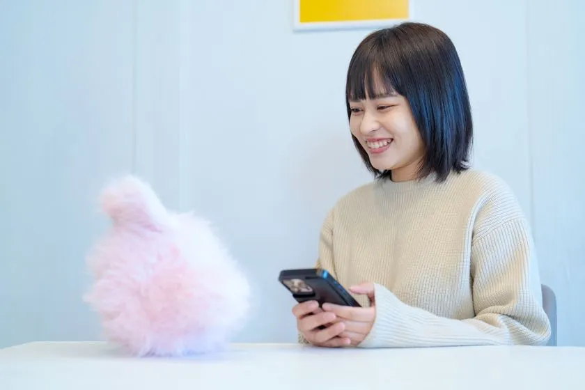 ピンク色のKOTTIぬいぐるみをテーブルに置き、スマートフォンを手に笑顔で操作する女性のシーン。スマホ連携・アプリ操作時の使用イメージを示す生活シーン写真