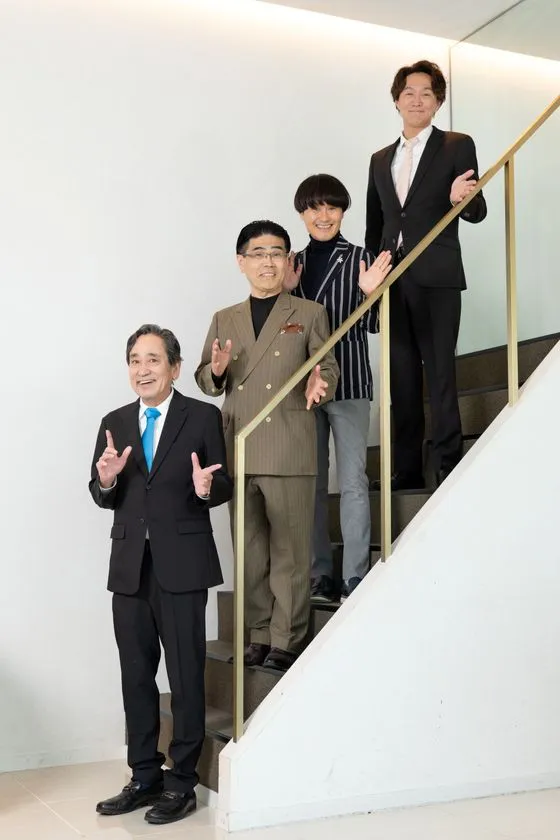 階段上に並んで手を振るポーズの4名全員集合写真。黒スーツの男性・ダブルスーツの男性・ストライプ衣装の人物・スーツの若い男性が縦並びに整列。