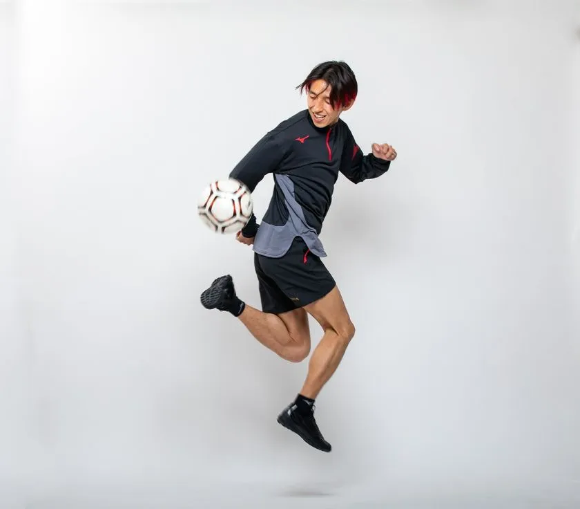 ドリブルデザイナー岡部将和氏がミズノ製黒ジャージ上下を着用しジャンプしながらサッカーボールをトラップする全身スタジオ撮影ショット。正面寄りのアングルで動きと表情が明確に写っている