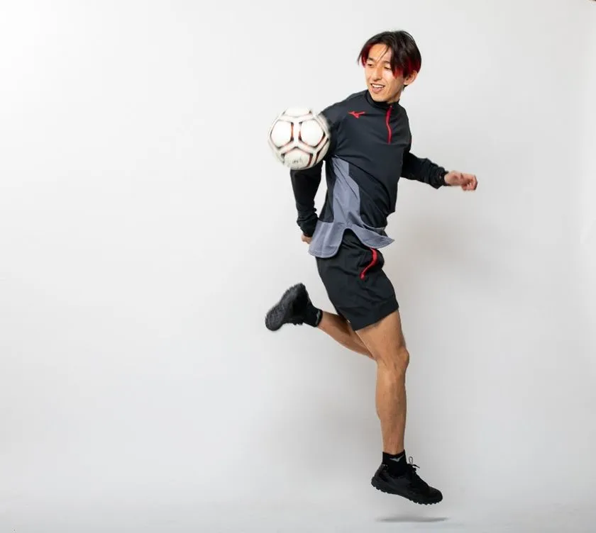 岡部将和氏が同じミズノ製黒ジャージでサッカーボールをインサイドでコントロールしながら空中に浮く全身スタジオ撮影ショット。画像1より左寄り構図で全身バランスと技術動作が見やすい角度