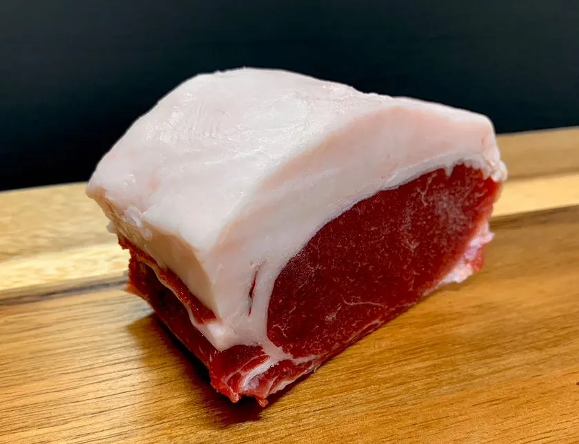 木製まな板の上に置かれた脂付きジビエ肉の塊。厚めの白い脂身と濃い赤身が断面で確認でき、イノシシまたはシカのブロック肉と見られる