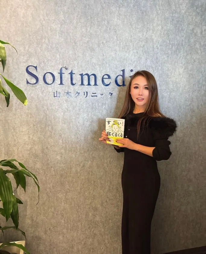 著者・山本江示子が黒ワンピース姿でSoftmedi山本クリニックのロゴ前に立ち、書籍『すごい!耳くるくる』を手に持つポートレート写真