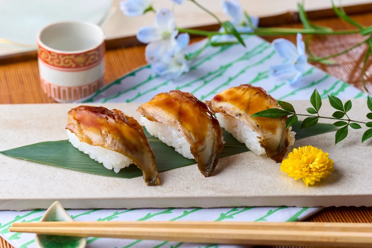 タレを塗って焼き上げた穴子の握り寿司3貫を石板の上に笹の葉を敷いて盛り付け、箸・菊花・青い小花で和の趣を演出した料理写真