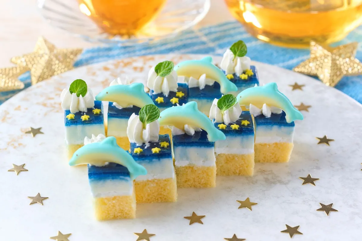 イルカのチョコ飾りとホイップクリーム・ミントを乗せた青×白×黄色のレイヤーゼリーケーキ（一口サイズ）が6個並び、テーブルには金色の星型コンフェッティとティーカップが添えられた夏季限定デザートの商品写真