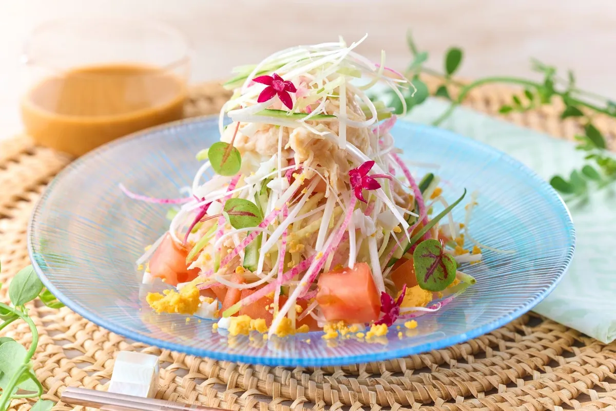 ほぐし蟹と千切り野菜を高く盛り付けた冷製サラダの単品アップ。青いガラス皿にトマト・スプラウト・食用花を添えた夏向け一皿