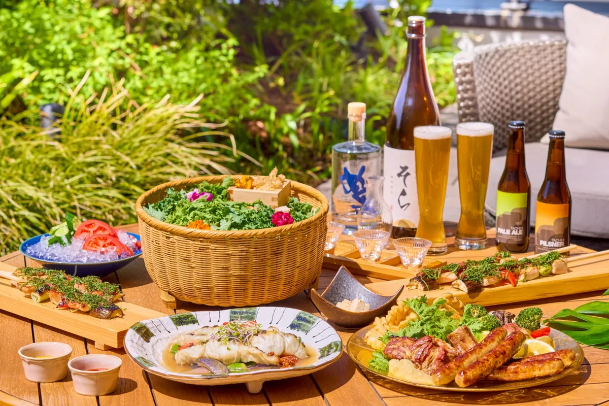 ソラノルーフトップバーの屋外テーブルに並ぶ夏メニュー全景。かごサラダ・魚料理・ソーセージ串焼き・野菜串を中心に、立川クラフトビール瓶・ペールエール・ピルスナー・日本酒ボトルが配置され複数品目を一覧できる料理スプレッドショット