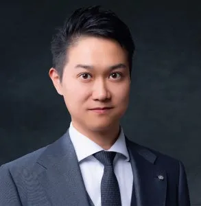 スーツ・ネクタイ姿の30代前後と思われる男性登壇者の単体プロフィール写真。ダークグレーの背景でビジネスフォーマルな上半身ショット
