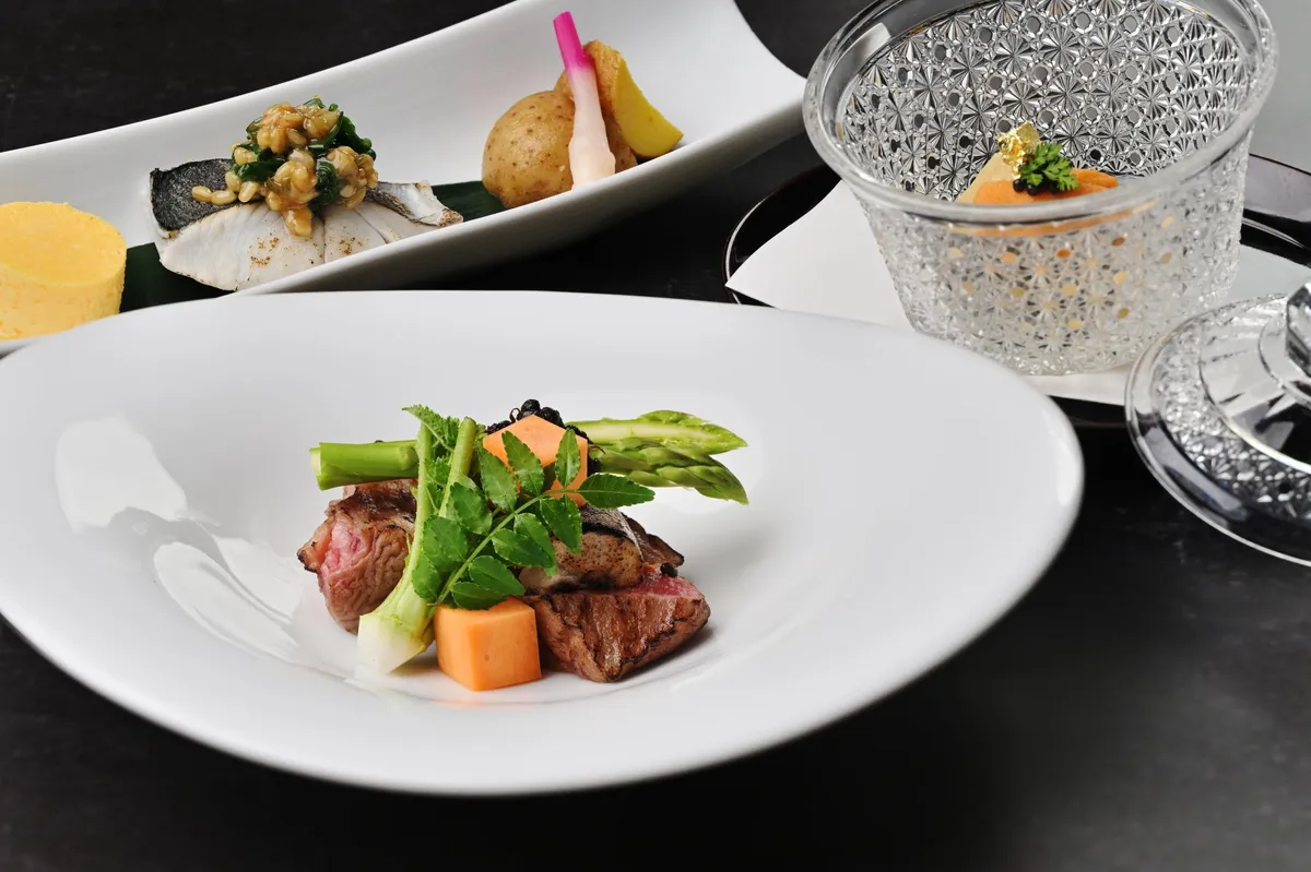 グランキュイジーヌ・コースの複数料理を俯瞰した盛り付け写真。手前の白皿に牛肉とアスパラ・人参・三つ葉を添えたメイン、奥に魚料理と野菜の前菜プレート、右にクリスタルガラス器の冷製スープが並ぶ