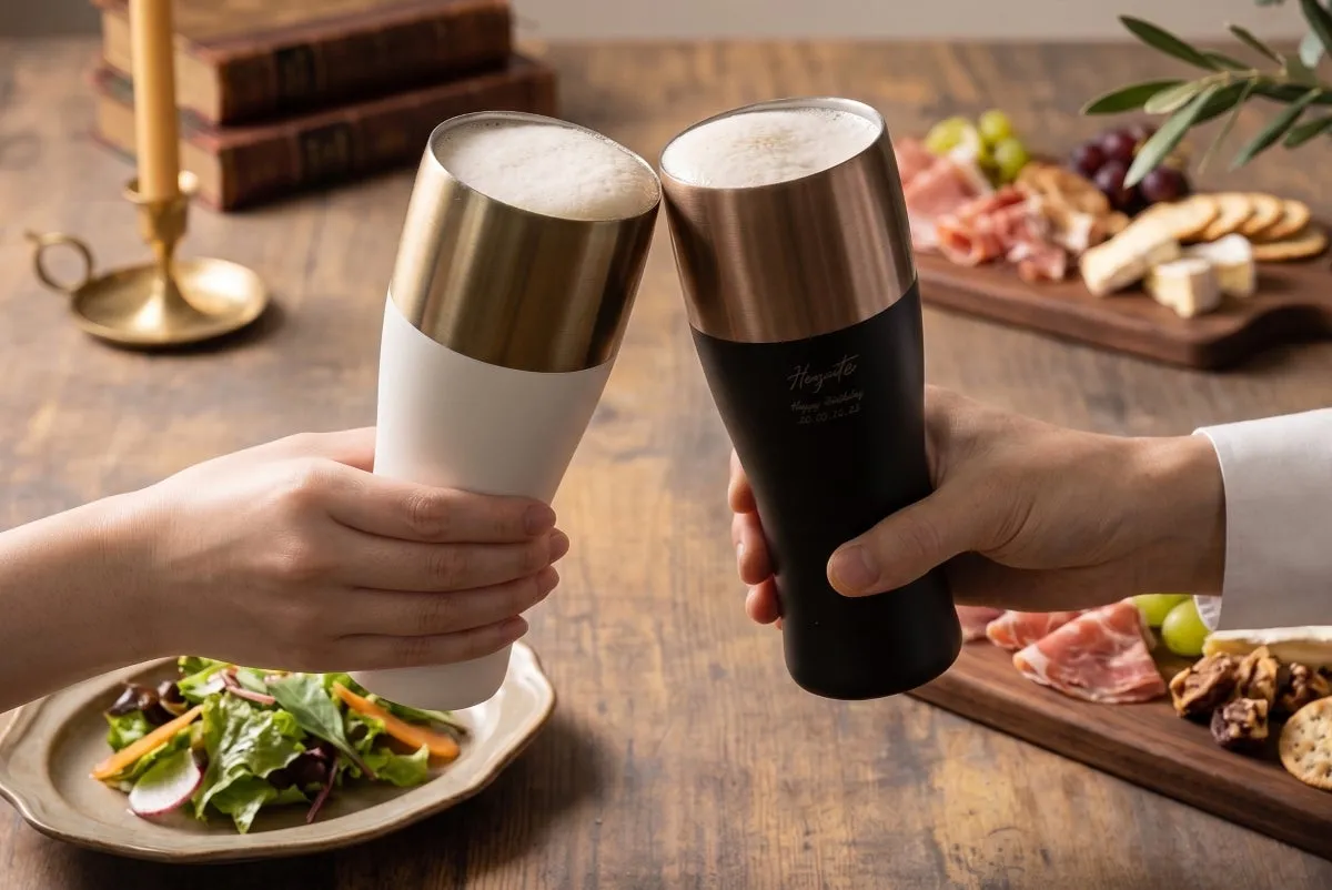 ホワイトとブラック2色の名入れ刻印タンブラーを両手で乾杯しているシーン。泡立つビール入り、背景にチーズ・ハムのグラスプレートとキャンドル