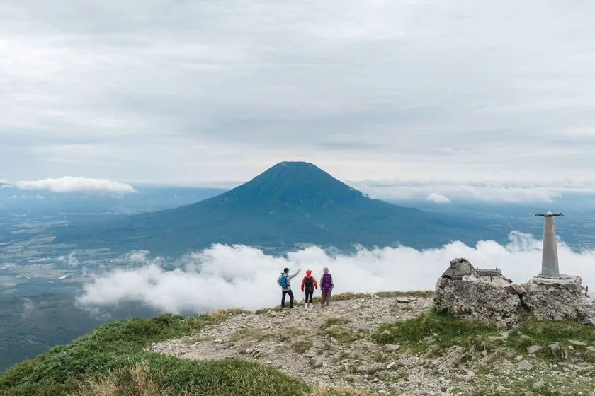 ニセコ連峰の山頂付近で3人のハイカーが羊蹄山を望む登山シーン。三角点標柱と雲海が広がる夏山の全景