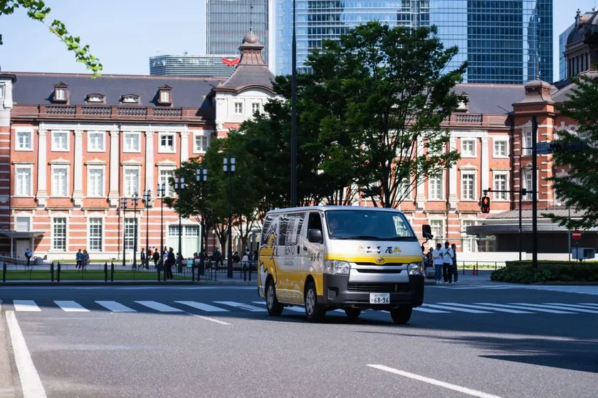 玉子屋のロゴと黄色帯デザインが入ったトヨタ・ハイエースの配達バンが東京駅丸の内口前の交差点を走行している配達車両ショット