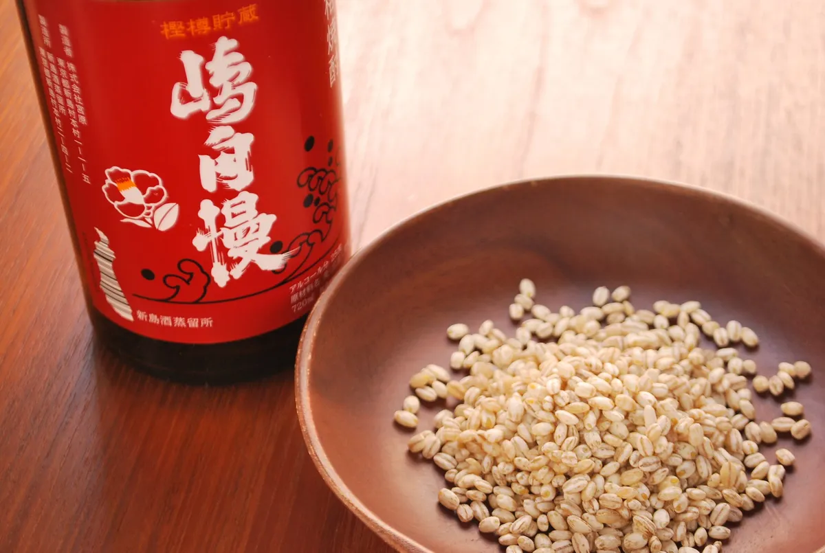 新島酒蒸留所の焼酎嶋自慢720ml赤ラベルボトルと、木製小皿に盛られた発酵原料の麦麹粒を並べた原料紹介ショット