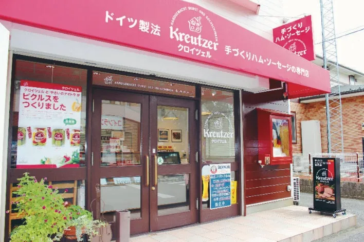 ドイツ製法手づくりハム・ソーセージ専門店クロイツェルの外観。赤い看板にKreutzerのロゴと店名が表示され、入口ドアや店頭のA型看板も確認できる