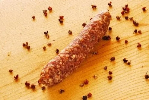 花椒（赤い実）を木製まな板に散らした中央に花椒入り焼酎麹発酵サラミ1本を置いた商品・原材料の組み合わせショット