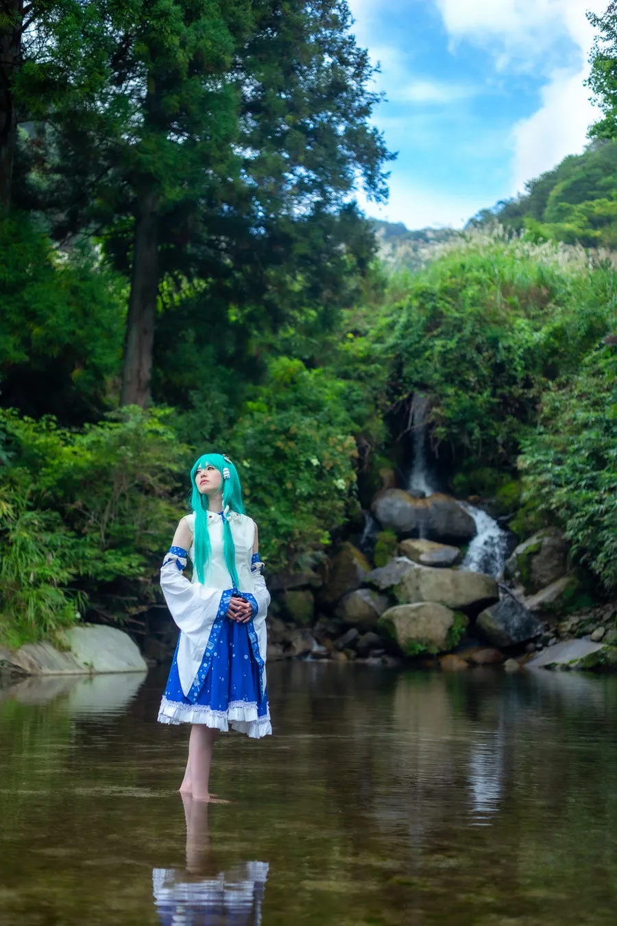 水色ロングヘアに白青ドレスのコスプレ人物が渓流の浅瀬に立ち滝を背景に上を向くポーズのロケ撮影カット。水面への反射と緑・青空の背景構成が明確。