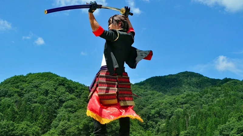 刀剣乱舞系の赤・黒・金の武者鎧コスプレ姿のキャラが青空と緑山を背景に刀を振り上げる後ろ姿アクションショット