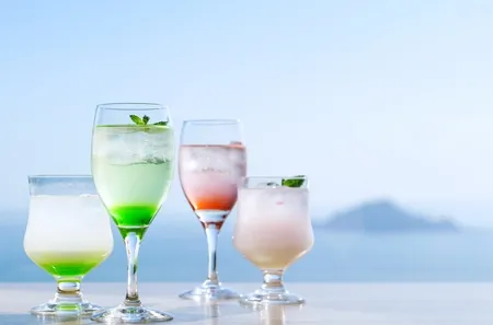 海を背景にテラス席に並ぶ4種のメロン系カクテル・ドリンク。緑・ピンク・白のグラスが並び伊良湖の海景色とともに撮影されたドリンクビジュアル