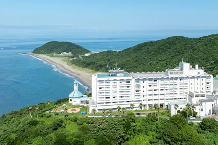 伊良湖オーシャンリゾートの白い高層ホテル建物と礼拝堂を緑の丘と海岸線・青い海を背景に俯瞰で捉えた外観全景写真