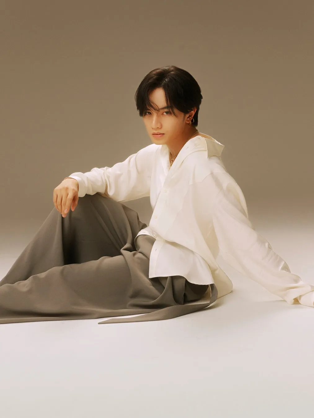 中島健人さんがグレーのワイドパンツと白シャツ姿でスタジオの床に座り、カメラを見つめるポートレート写真。ゴールドのネックレスとピアスを着用。