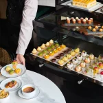 スタッフがワゴンを客席へ引き出しスイーツをサーブする場面。ワゴン上段にケーキ・カヌレ・シュー、中段にフルーツタルト・チョコカップ、下段にムース小瓶が並び、テーブルにはセイボリー皿も置かれる