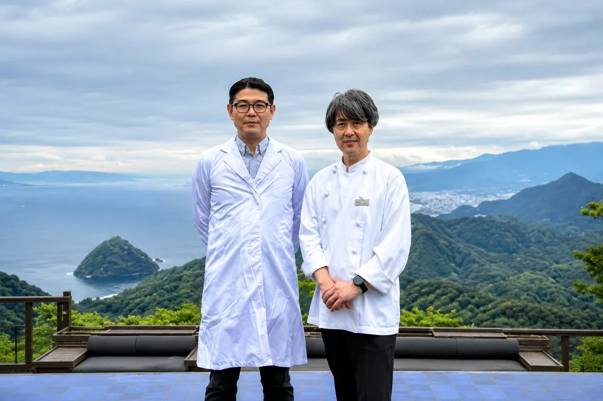 白衣着用の研究者風の男性とシェフコート姿の男性2名が、伊豆の山並みと海を望む高台テラスで並んで立つ連携先担当者の対面写真