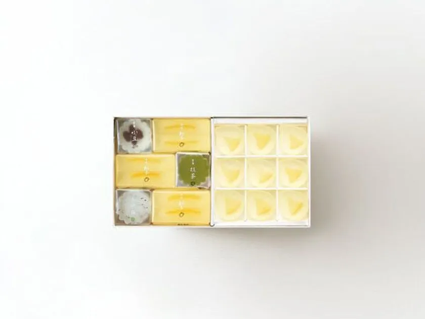 白い紙箱に収めた永楽屋の和菓子詰め合わせ。左区画に小豆・抹茶・桜風味の羊羹と練り切り8個、右区画に淡黄色の琥珀糖（レモンの雫）9個を3×3格子配置した俯瞰ショット。