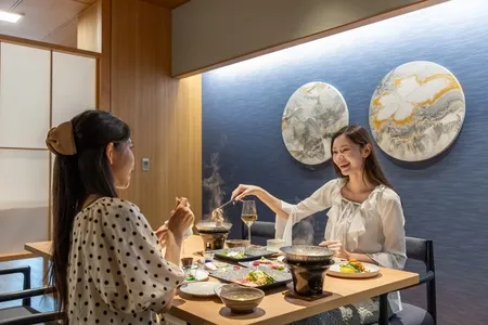 和モダンな個室ダイニングで女性2人が食事を楽しむ場面。テーブル上に多皿の日本料理が並び、壁面に大理石調の丸型アート2枚が飾られている