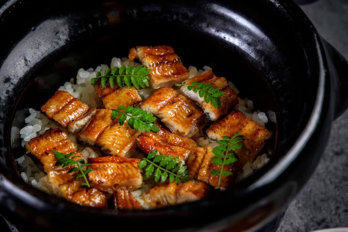 黒い土鍋に盛られた鰻の土鍋ご飯。タレで照り焼きにされた鰻の切り身が白米の上に並び、山椒の葉が数枚あしらわれている料理写真