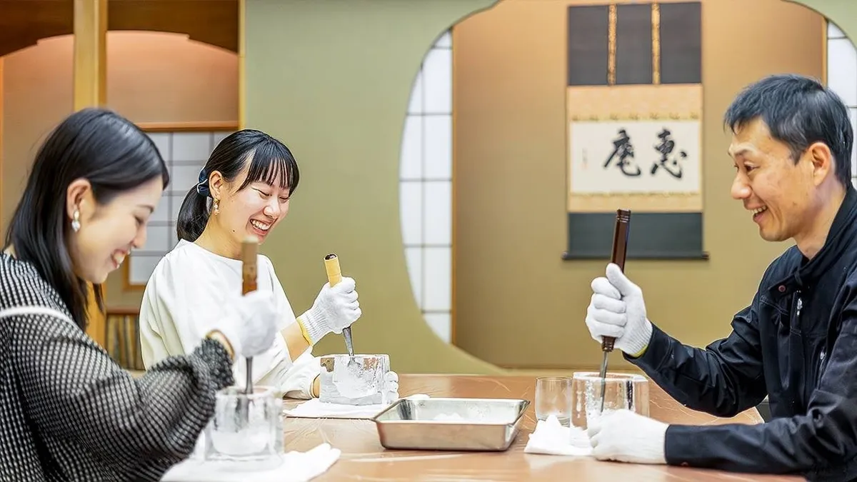 和室の丸窓と掛け軸を背景に3名の参加者が白手袋をはめ氷彫刻用ノミで氷を削る体験プログラムのシーン