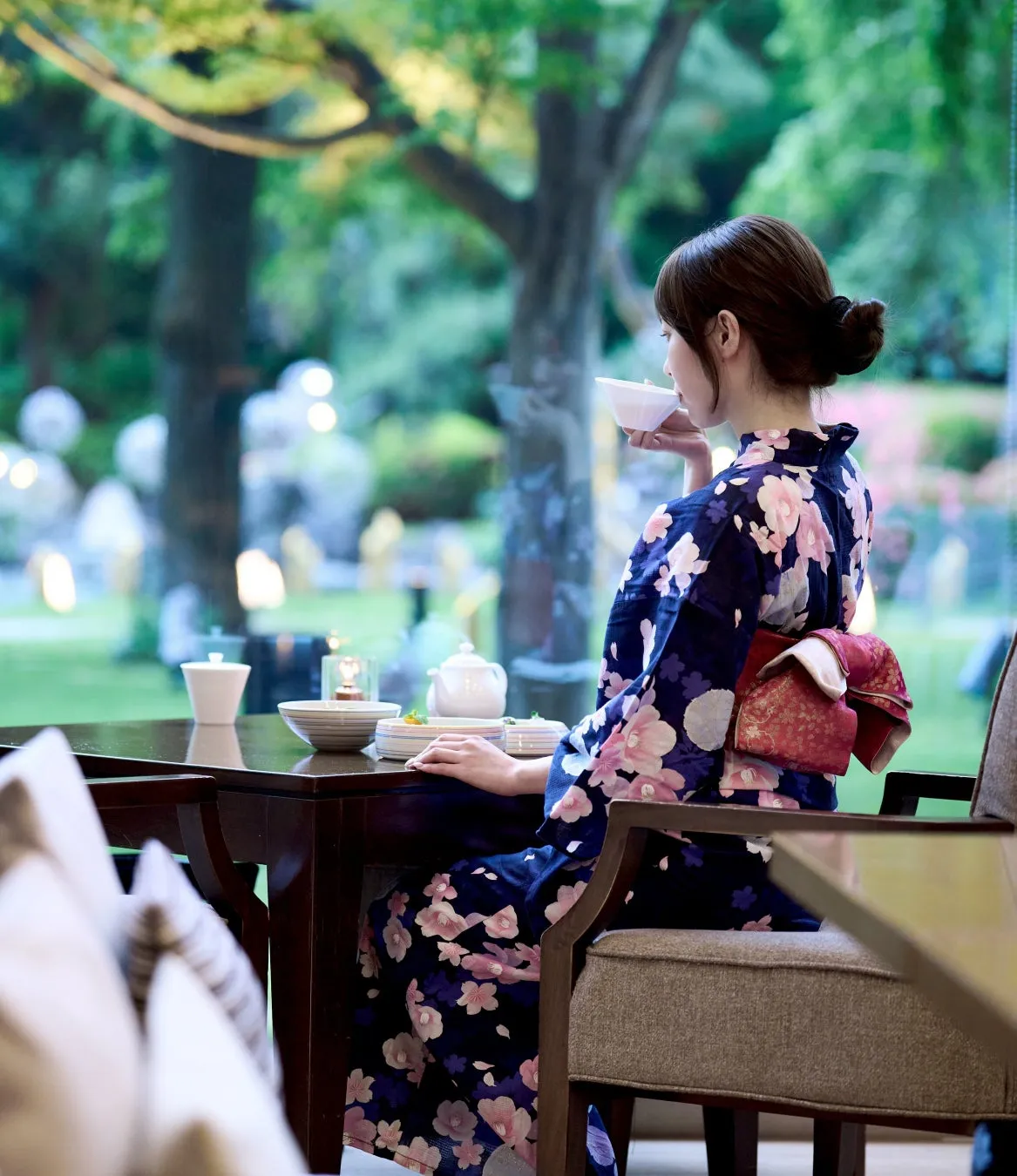 紺地桜柄浴衣に赤帯を締めた女性が日本庭園に面した窓際の椅子に座り白い茶碗を口に運ぶ横顔ショット