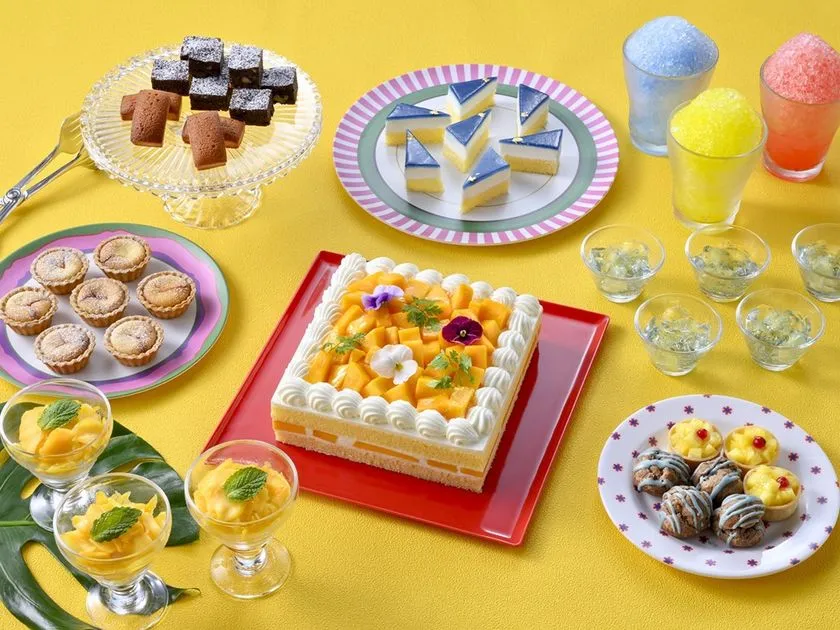 夏のスイーツビュッフェ品揃えを上から撮影した俯瞰写真。マンゴーケーキ・チョコブラウニー・タルト・かき氷ドリンク・パンナコッタなど10種以上のデザートを黄色背景に配置